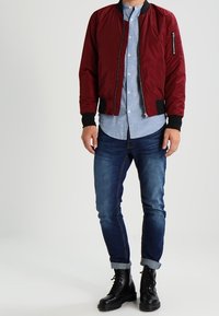 Bordeauxfarbene Bomberjacke mit schwarzen Rippbündchen und einer mit Reißverschluss versehenen Brusttasche, getragen über einem hellblauen Hemd mit Knopfleiste und dunklen Jeans, kombiniert mit schwarzen Stiefeln.