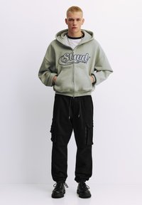 Hanorac gri cu glugă, logo, buzunare frontale și închidere cu fermoar, asortat cu pantaloni cargo negri cu buzunare și manșete la glezne. Pantofi negri.