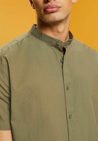 Close-up van een persoon die een olijfgroen mandarin kraag shirt met knopen aan de voorkant draagt, tegen een mosterdgele achtergrond.