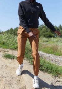 Schwarze Fleece-Zip-up-Jacke mit Logo, braune taillierte Cargo-Hosen und weiße Sportschuhe, vor einer natürlichen Outdoor-Kulisse.