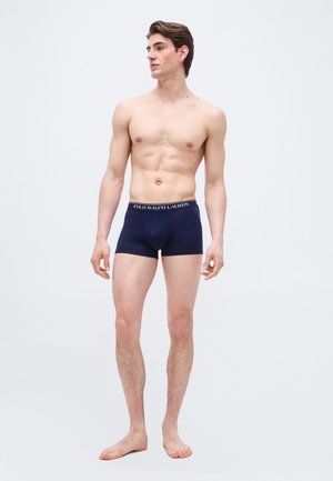 Νεαρός άνδρας, ξυπόλητος, φοράει μπλε σκούρα boxer briefs Polo Ralph Lauren, κοιτάζοντας προς τα αριστερά του σε απλό ανοιχτό φόντο.