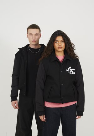 Giacca nera con corpo in lana e maniche nere, dotata di un logo e due tasche frontali. Il modello indossa una camicia rosa sotto.