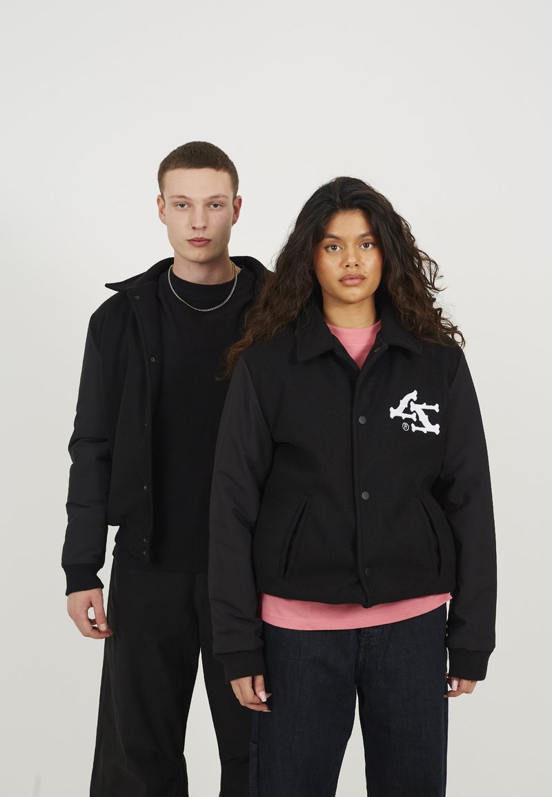 Zwarte jas met een wollen lijf en zwarte mouwen, voorzien van een logo en twee voorzakken. Het model draagt eronder een roze shirt.