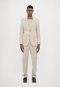 Completo beige chiaro con giacca a due bottoni e pantaloni a taglio dritto, abbinato a una camicia verde chiaro e scarpe nere. Presenta una spilla bianca all'occhiello.