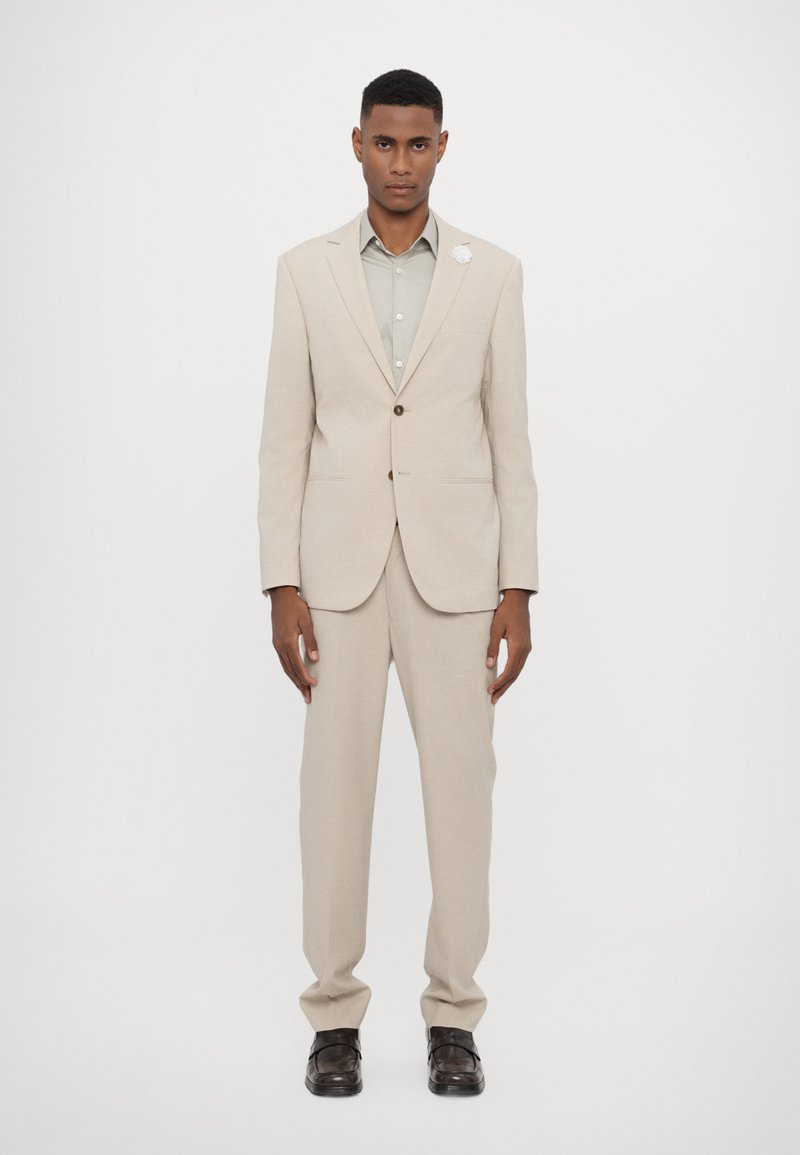 Completo beige chiaro con giacca a due bottoni e pantaloni a taglio dritto, abbinato a una camicia verde chiaro e scarpe nere. Presenta una spilla bianca all'occhiello.