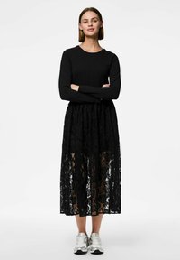 Robe noire à manches longues avec une jupe en dentelle noire transparente ornée de motifs floraux, assortie à des baskets argentées. Haut lisse avec un col rond.