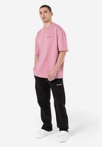 Tricou oversized roz deschis, cu mâneci scurte și un mic logo pe piept, asortat cu pantaloni negri și adidași albi.