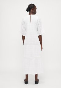 Rochie maxi albă cu mâneci scurte evazate, model texturat în carouri și dantelă decorativă. Dispune de o deschidere în formă de ochelt la spate.