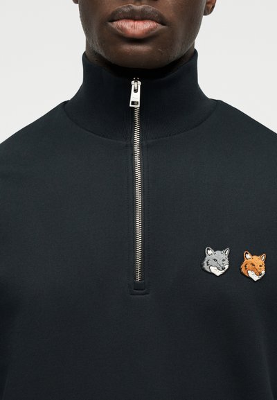 Κοντινό πλάνο προσώπου που φοράει μαύρο πουλόβερ με φερμουάρ και ψηλό γιακά, με μικρά κεντημένα γκρι και πορτοκαλί κεφάλια αλεπούδων στο στήθος.