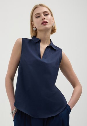 Donna che indossa una blusa senza maniche color navy con colletto, mani nelle tasche, orecchini a cerchio argentati e un bracciale spesso argentato su uno sfondo semplice.