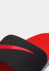 Μαύρο σανδάλι slide με μια κόκκινη κυρτή ραβδωση στο λουρί και μια υφή κόκκινης σόλας σε λευκό φόντο.