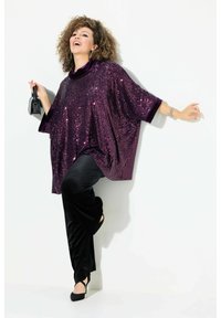 Top oversized in paillettes viola con pantaloni in velluto nero e tacchi neri. Una piccola borsa nera è tenuta in una mano.