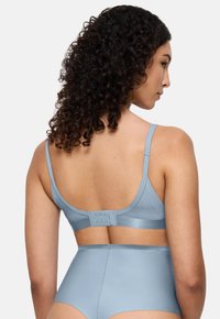 Woman with curly dark hair wearing light blue bra with adjustable straps and high-waisted underwear, shown from the back against a white background.