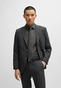 Costume gris charbon avec une cravate texturée, design ajusté et motifs à carreaux discrets. Veste et pantalon sur mesure, avec une ceinture noire élégante.