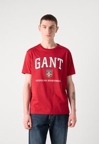 Tricou roșu din bumbac cu text îndrăzneț "GANT" și logo-ul unui scut. Guler rotund și mâneci scurte, asociat cu blugi din denim de culoare albastru închis.