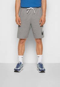 Man med grå dragbandsshorts med stort logo på vänster ben, vita Nike-strumpor och blå och grå sneakers, stående mot en vit vägg.