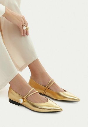 Füße in spitzen goldenen flachen Schuhen mit doppelten Riemen und großen Perlenknöpfen, kombiniert mit hellbeigen Hosen und einer Hand, die auf dem Knie ruht.