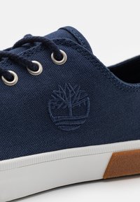 Baskets en toile bleue avec une semelle en caoutchouc blanche, dotées d'œillets argentés et d'un logo d'arbre brodé. La texture est lisse, avec des détails cousus.