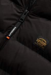 Schwarze Daunenjacke mit gesteppten Abschnitten, ausgestattet mit einem Frontreißverschluss und einem Gewebelabel in Gold und Schwarz, auf dem "SUPERDRY" steht.