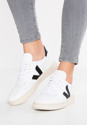 Baskets blanches à lacets avec des accents noirs, un design perforé sur l'orteil, un matériau texturé et une semelle en caoutchouc beige.