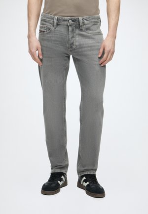 Homme portant un jean gris délavé et des baskets noires avec des accents blancs, debout devant un fond clair uni.
