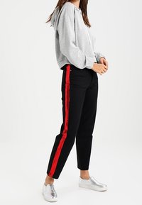 Donna che indossa pantaloni neri con strisce rosse ai lati, una felpa grigia corta e scarpe slip-on argentate, in piedi su uno sfondo bianco.