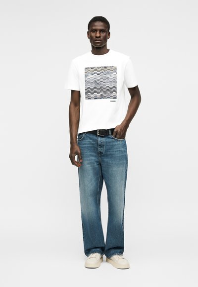 Άνδρας που στέκεται με το πρόσωπο προς τα εμπρός, φοράει λευκό μπλουζάκι με κυματιστό μοτίβο, μπλε τζιν, μαύρη ζώνη και λευκά αθλητικά παπούτσια σε λευκό φόντο.