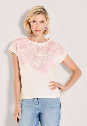 Frau mit kurzen blonden Haaren trägt ein hellrosa Oberteil mit floralem Muster und hellblaue Jeans, posiert mit einer Hand in der Hüfte.