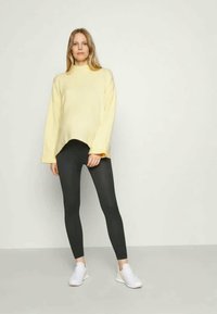 Suéter amarelo de malha com gola alta e mangas oversized, combinado com leggings pretas e ténis brancos, numa tela neutra.