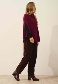 Vinröd stickad poncho med sidoflikar, bärs över en svart långärmad topp. Bruna kordtäckta byxor och vinröda klackade ankelstövlar fulländar looken.