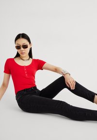 Top curto de manga curta em vermelho com textura em nervuras, apresentando detalhes em botão, combinado com jeans skinny pretos de cintura alta. Os acessórios incluem pulseiras prateadas e óculos de sol.