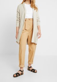 Lång beige kavaj, vit åtsittande skjorta, skräddarsydda ljusbruna byxor med slipsbälte och svarta sandaler med remmar och korksula.