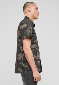 Tričko s krátkym rukávom v maskáčovom vzore v odtieňoch zelenej, sivej a modrej. Vyrobené z ľahkej tkaniny, s gombíkmi na prednej strane a golierom.