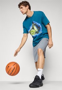 Teal grafisk t-shirt med basketball-design, grå træningsshorts og sorte sneakers. Holder en basketball, står på en hvid baggrund.
