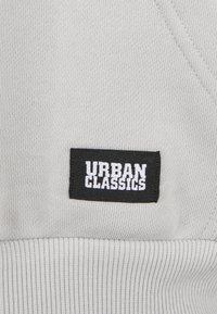Svart rektangulär etikett med vit text "URBAN CLASSICS" sydd på ljusgrå ribbad tyg.