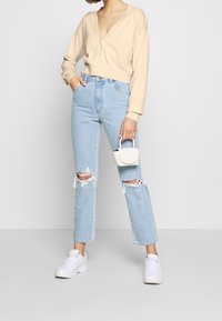 Beige kort cardigan med djup V-ringning, matchad med ljusblå slitna jeans med revor, och vita sneakers, hållandes en liten vit väska.