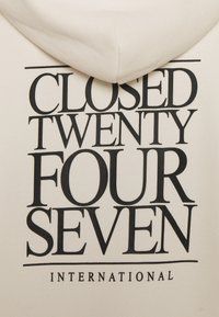 Sweat à capuche crème avec un texte noir audacieux indiquant "CLOSED TWENTY FOUR SEVEN INTERNATIONAL" en différentes tailles de police au dos.