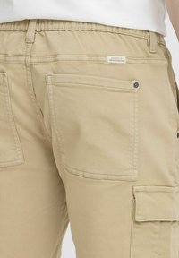 Pantalones cargo beige con cinturilla elástica, que cuentan con múltiples bolsillos y una textura de tela suave. La etiqueta dice "Dedicado a la Perfección del Denim".