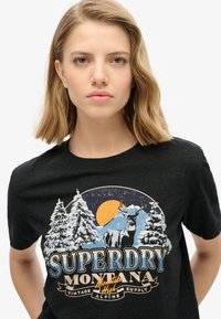 T-shirt en coton noir avec un graphisme de montagne et de forêt, incluant un élan, un soleil et le texte "SUPERDRY MONTANA" dans plusieurs couleurs.
