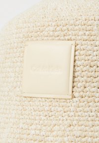 Tejido de color beige con superficie texturizada; cuenta con un parche de cuero de color beige claro etiquetado "Calvin Klein". Forma redonda, diseño casual.