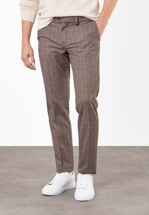 Man draagt bruine geruite broek en witte sneakers, staat op een grijze vloer met één hand in zijn zak.