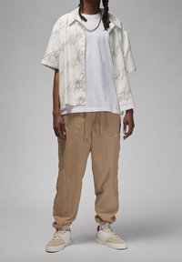 Kortärmad vit skjorta med guldscriptdesign, kombinerad med beiga tapered byxor och vita sneakers med röda accenter. Lagerhalsband synligt.