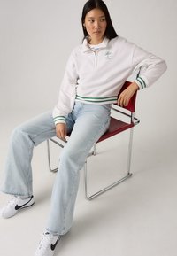 White fleece pullover with green stripes at cuffs, zip neck detail, and text logo. Light blue high-waisted jeans and white sneakers.