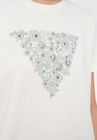 Camiseta blanca de algodón con un diseño de encaje triangular de color azul claro adornado con cuentas decorativas y patrones florales.