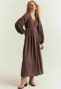Maxi sukienka w stonowanej mauve, z dekoltem w kształcie V, bufkami i subtelnymi haftowanymi detalami. Stylizowana z brązowymi botkami za kostkę.