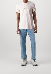 Camiseta de algodón rosa claro con el logo "GUESS" en rojo, combinada con jeans rectos azul claro y zapatillas plateadas con acentos en neón.
