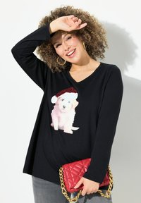 Schwarzes Langarm-T-Shirt mit V-Ausschnitt, das ein Grafikbild eines weißen und rosa Welpen zeigt, der einen rot-weißen Weihnachtsmannhut trägt. Graue Jeans und eine rote gesteppte Handtasche.