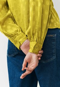 Gelbes Hemd aus strukturiertem Stoff mit großen Punkten, ausgestattet mit geknöpften Manschetten, kombiniert mit dunkelblauen Jeans.