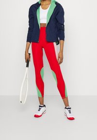 Červené legíny se zelenými akcenty a růžovými bočními panely, spárované s námořnickou modrou zipovou bundou se zeleným vnitřkem. Drženy bílé sportovní boty.