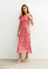Robe midi florale rouge avec des manches courtes volantées, un corsage ajusté et une jupe fluide. Associée à des talons blancs à lanières et à un petit sac à main.